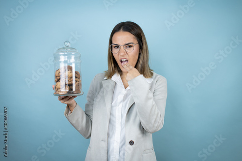 Young beautiful woman holding chocolate chips cookies jar over isolated blue background disgusted with her hand inside her mouth photo