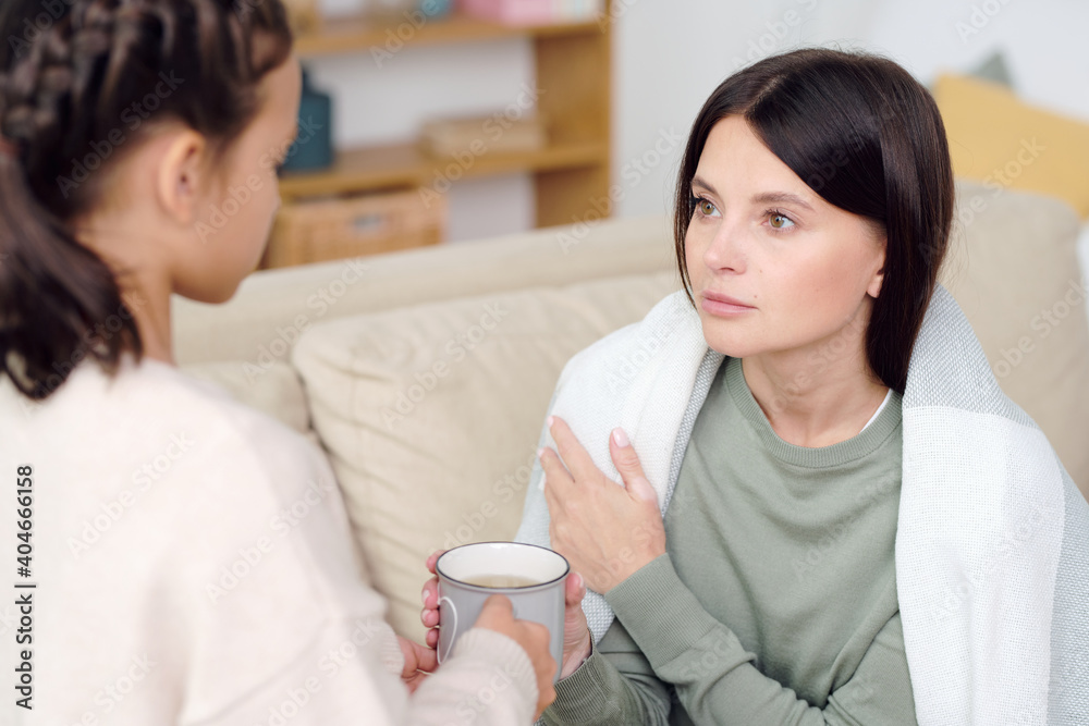 Young brunette sick female wrapped into warm plaid looking at her daughter Young brunette sick female wrapped into warm plaid looking at her daughter