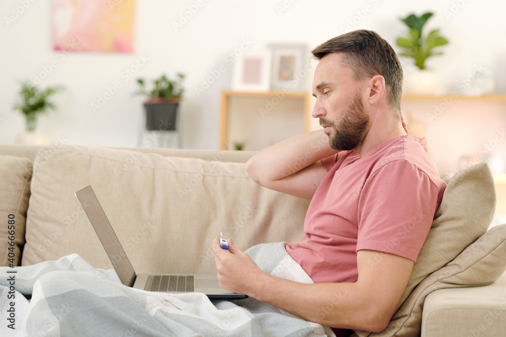 Side view of young sick man with thermometer sitting on couch under blanket Side view of young sick man with thermometer sitting on couch under blanket