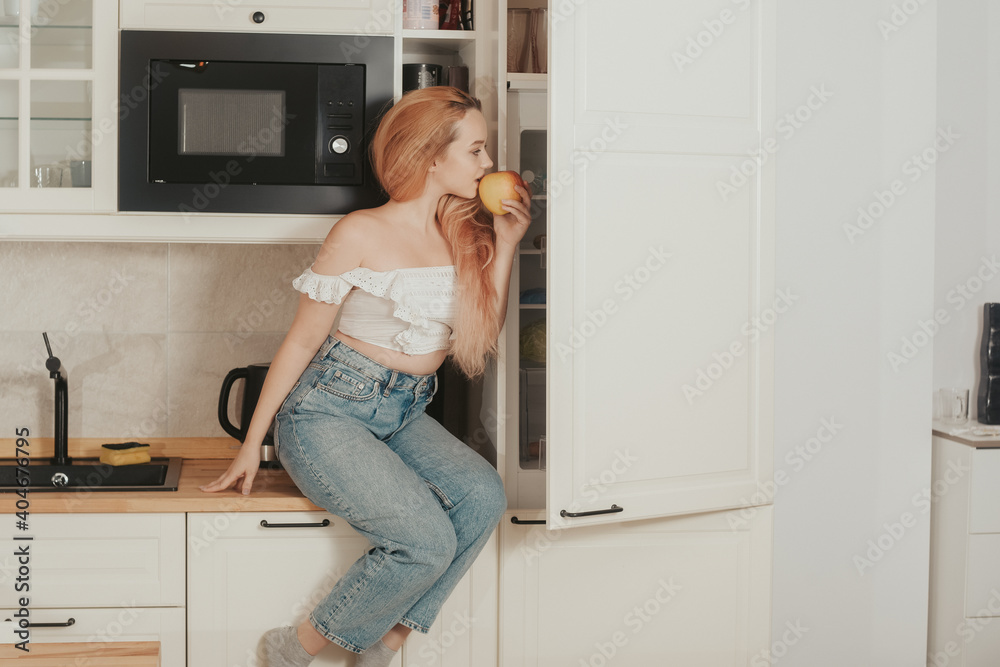 A woman is sitting at home in the kitchen near the refrigerator. A girl with a good figure looks in the refrigerator, healthy food, diet. The girl sits on the table and eats an apple A woman is sitting at home in the kitchen near the refrigerator. A girl with a good figure looks in the refrigerator, healthy food, diet. The girl sits on the table and eats an apple