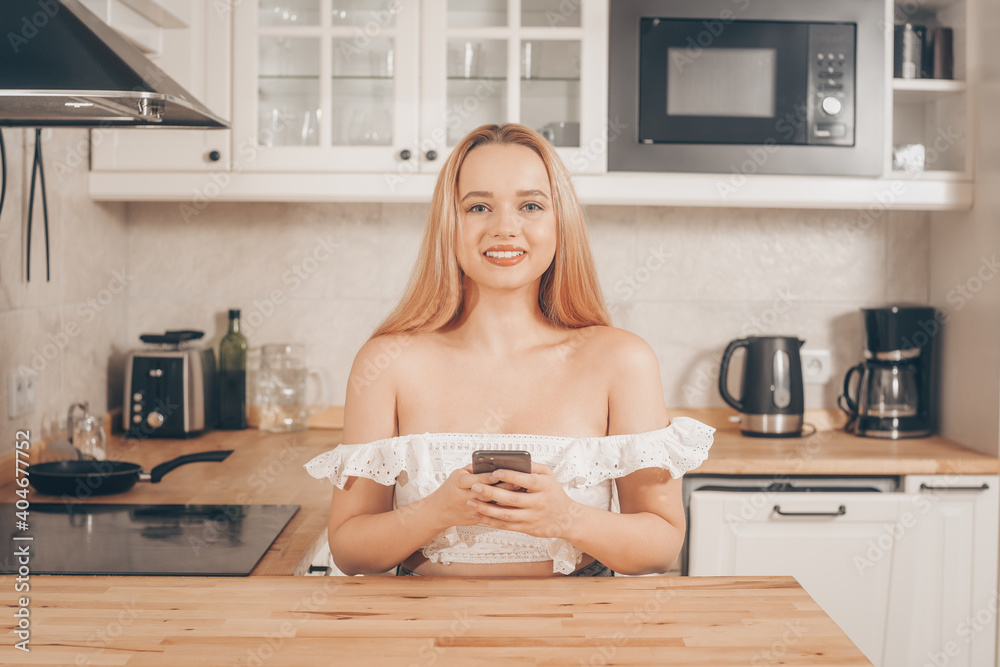 Beautiful woman sits at a table on the background of the kitchen at home. Girl with a smart smart phone in her hands. The blonde writes a message or orders takeaway. Online delivery Beautiful woman sits at a table on the background of the kitchen at home. Girl with a smart smart phone in her hands. The blonde writes a message or orders takeaway. Online delivery