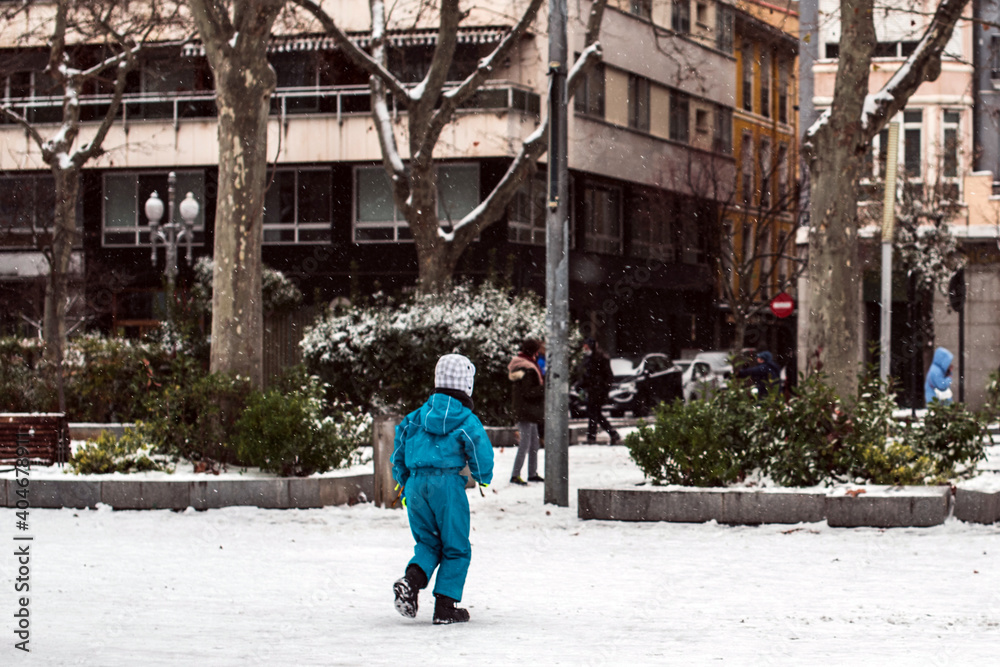 Niña pequeña vistiendo un mono azul de invierno como abrigo y un gorro de lana caminando sola en una calle cubierta de nieve en el centro de la ciudad Niña pequeña vistiendo un mono azul de invierno como abrigo y un gorro de lana caminando sola en una calle cubierta de nieve en el centro de la ciudad