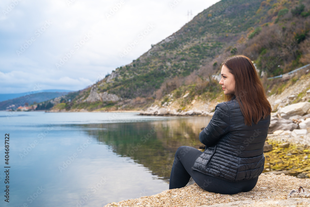 Young brunette woman in jacket sits and reflects on rocky shore of calm sea against background of mountains in peaceful atmosphere. Idyllic walk alone.