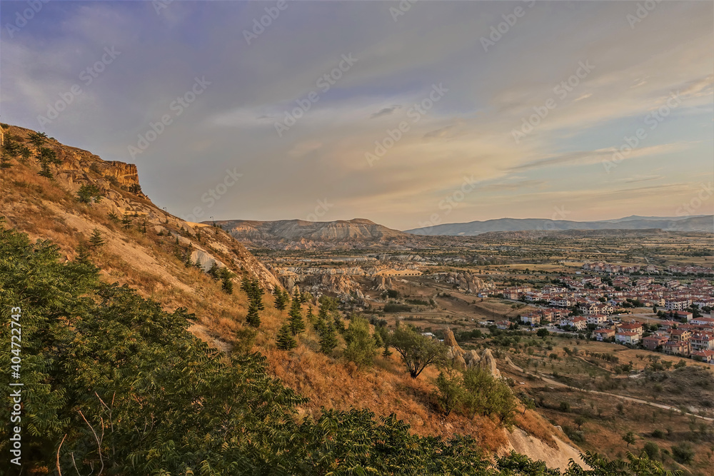 Cappadocia landscape at golden hour. In the foreground is a mountain slope with coniferous and deciduous trees. In the valley you can see bizarre rocks and a village. Clouds in the blue sky. Turkey