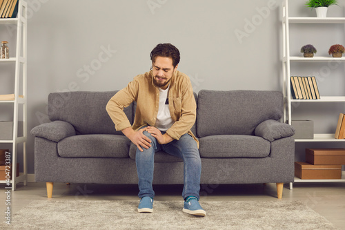 фотографія Man sitting on couch at home, unable to stand up, touching his hurt knee with grimace of pain