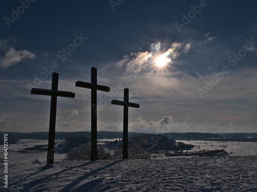 Three Christian crosses on the top of Kornbühl hill near Burladingen, Germany in low mountain range Swabian Alb with beautiful winter landscape and backlight of bright afternoon sun on cloudy sky. photo