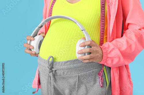 Pregnant woman putting headphones on her belly against color background