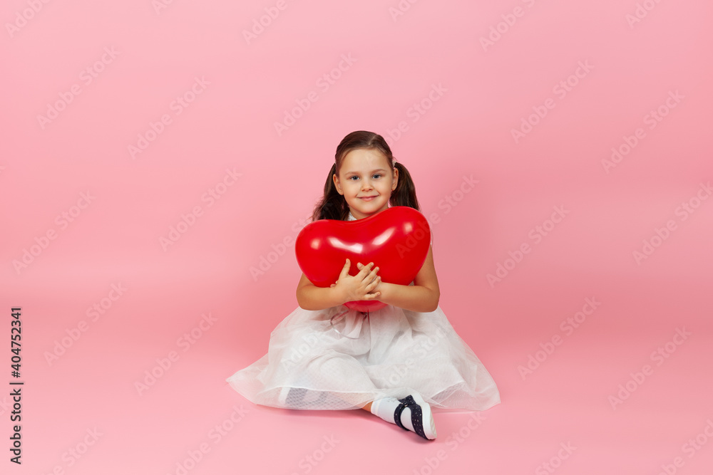 studio portrait of a nice, sweet girl in a white dress hugging a red heart-shaped balloon and sitting on the floor , isolated on a pink background. studio portrait of a nice, sweet girl in a white dress hugging a red heart-shaped balloon and sitting on the floor , isolated on a pink background.