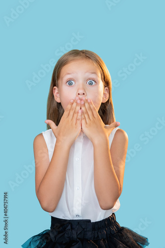 Shocked kid. Back to school. Unexpected news. Portrait of surprised overwhelmed cute little girl in formal outfit covering mouth with hands isolated on blue background.