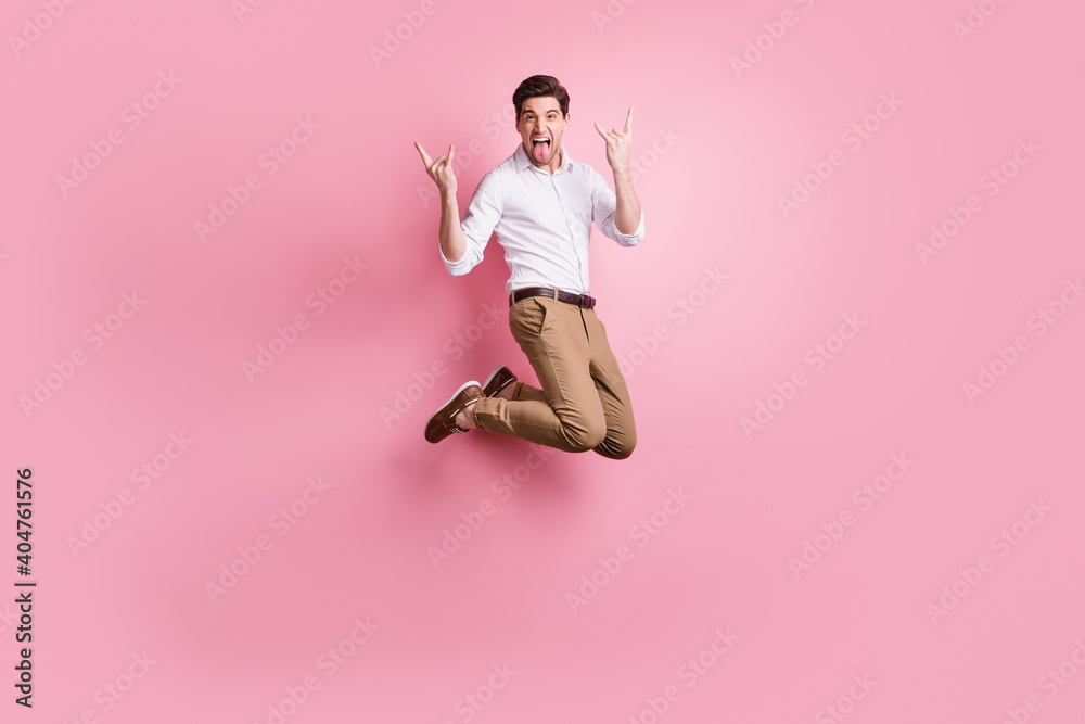 Full size photo of optimistic guy jump show rock sign wear white shirt trousers sneakers isolated on pink color background Full size photo of optimistic guy jump show rock sign wear white shirt trousers sneakers isolated on pink color background