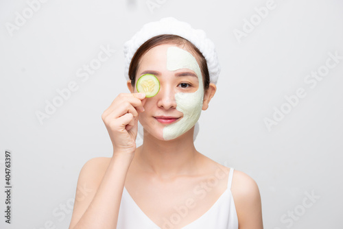 Beautiful young Asian woman with clay mask holding orange cucumber covering eye over white background.