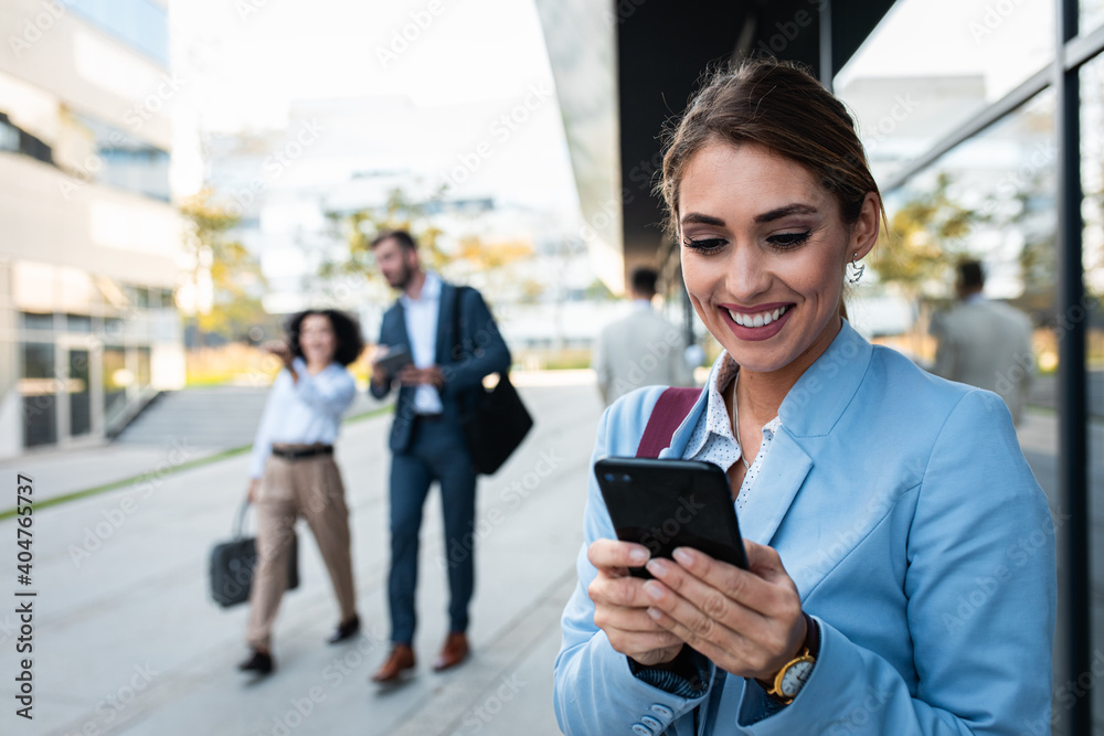 Portrait of smiling businesswoman standing in front of modern office buildings looking at phone.	