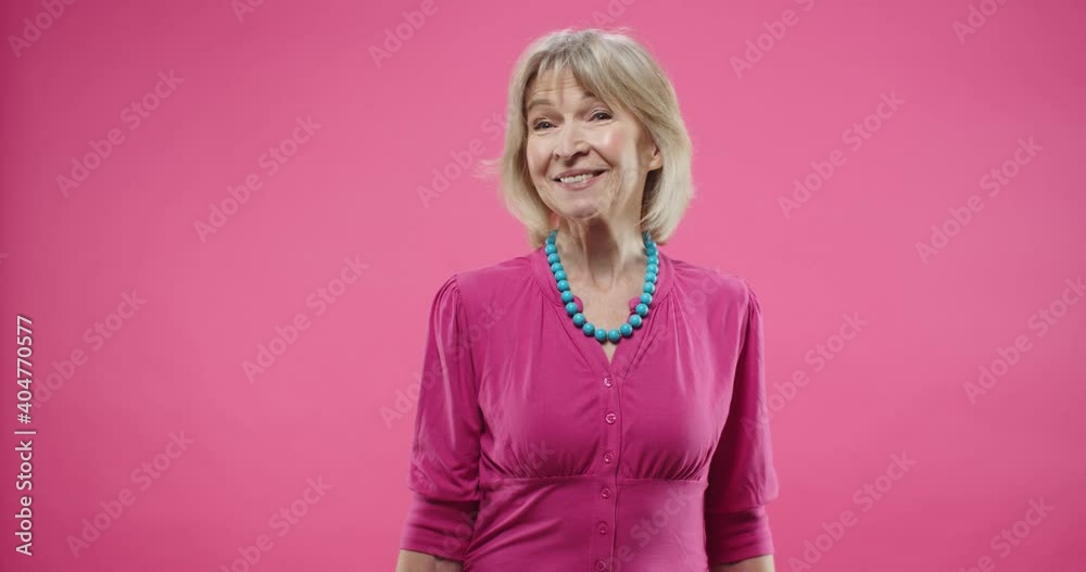Portrait of senior cheerful Caucasian pretty woman standing on pink background in good mood, smiling and looking at camera feeling pleased and happy. Beautiful old female in studio with smile on face