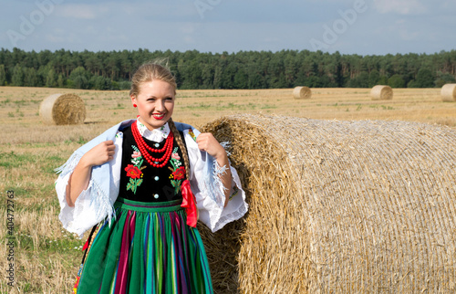 A pretty happy young girl dressed in Polish traditional national folk dress standing in the field on a harvest straw bales under a blue sky