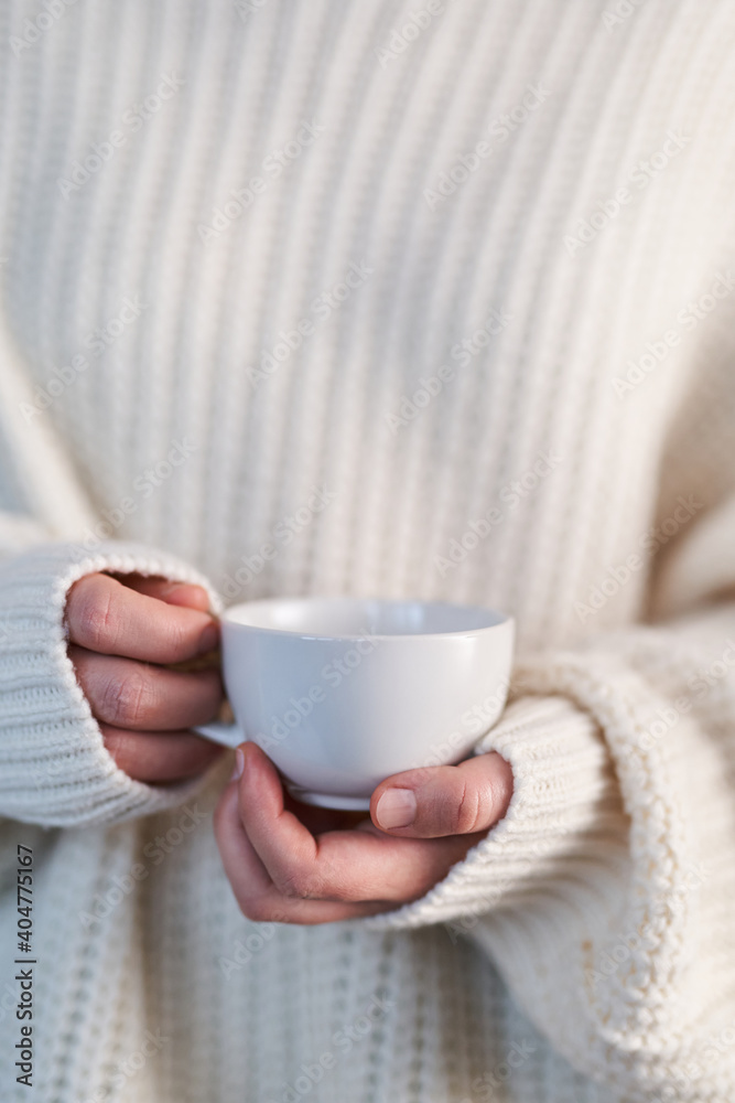 Female hands in beige sweater holding cup with hot drink. Close up white mug in female hands. Concept o winter comfort, morning time, warmly feeling
