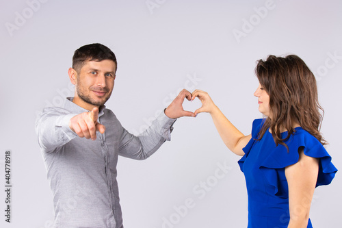 The couple is holding a heart shape from the hands. The man points his index finger at the camera. Gray background.