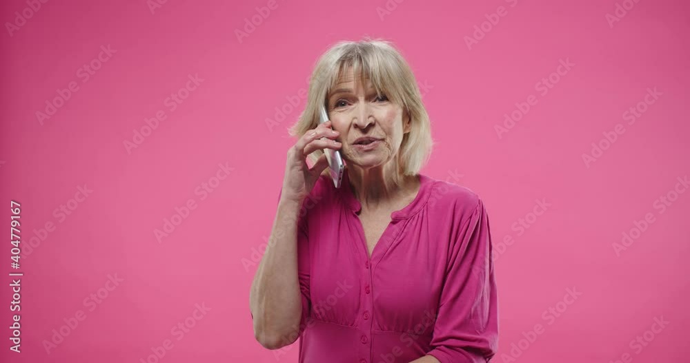 Close up portrait of cheerful senior Caucasian beautiful old woman with blonde hair standing isolated on rosy pink background in studio speaking calling on smartphone discussing something Close up portrait of cheerful senior Caucasian beautiful old woman with blonde hair standing isolated on rosy pink background in studio speaking calling on smartphone discussing something