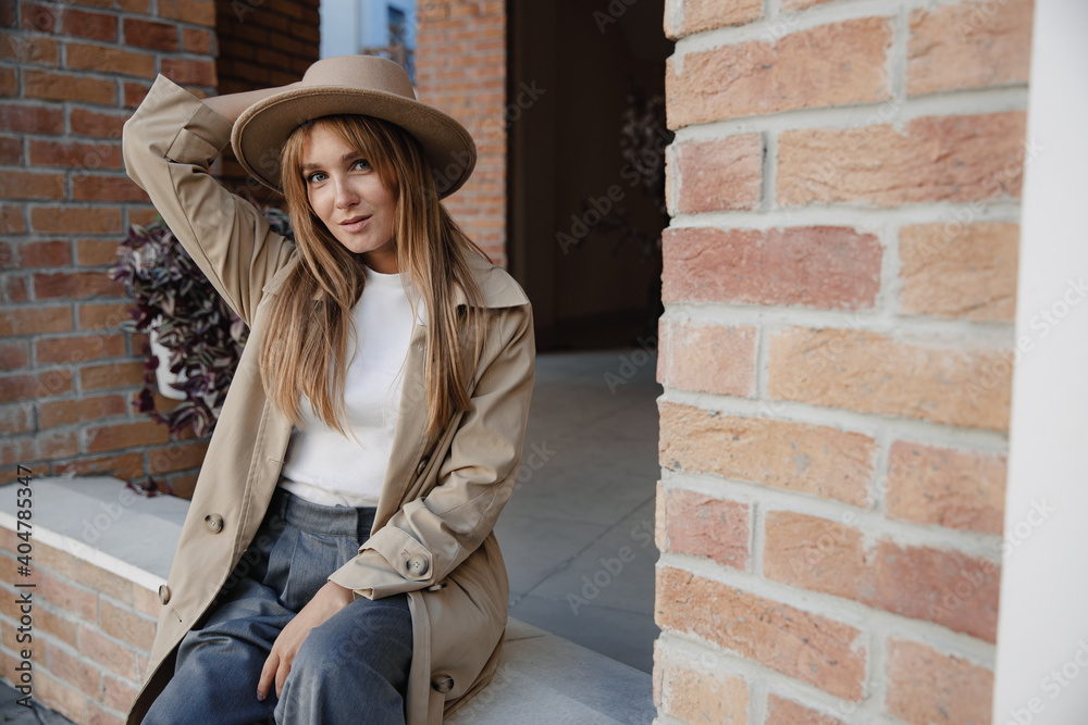 Fashion girl in an autumn trench coat and beige hat gently smiles and poses sits on the porch of brick house, stylish woman portrait.