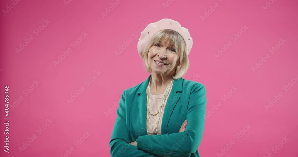 Portrait of happy smiling Caucasian senior old female in green jacket and beret standing isolated on pink background in positive mood looking at camera with happy face expression alone