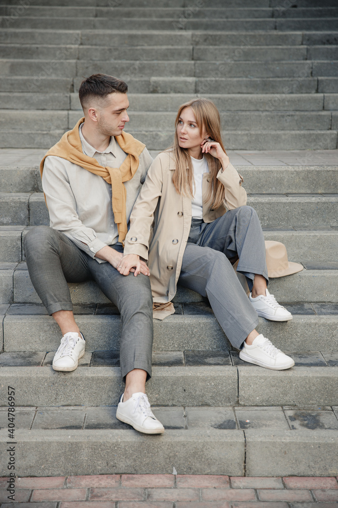 Portrait of couple in stylish autumn clothes are sitting on the stairs outside and looking at each other with love. Concept romantic date during quarantine and self-isolation