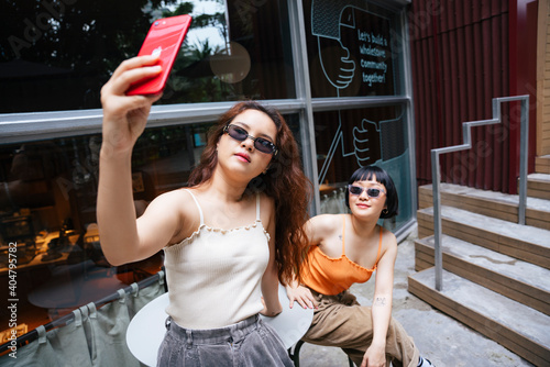 Portrait of two asian women wear sunglasses taking selfie with smartphone in cafe outdoors.