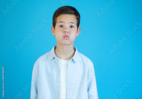 Beautiful kid boy wearing casual shirt standing over isolated blue background puffing cheeks with funny face. Mouth inflated with air, crazy expression.