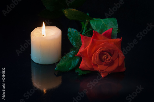 Red rose flower with water drops and white burning candle on black background with reflection .