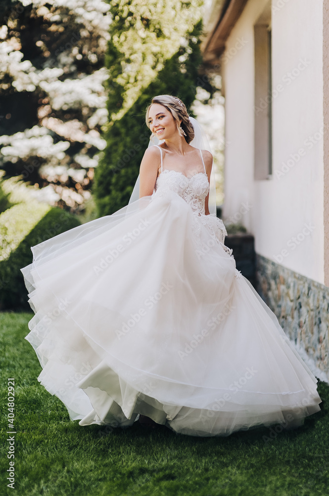 Fototapeta premium A beautiful, cheerful, happy, smiling blonde bride in a white lace dress is spinning and dancing in the park on the green grass against the background of the hotel building. Wedding portrait.