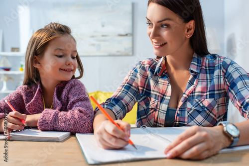 Smiling mother looking at daughter while writing in copy book at desk on blurred background
