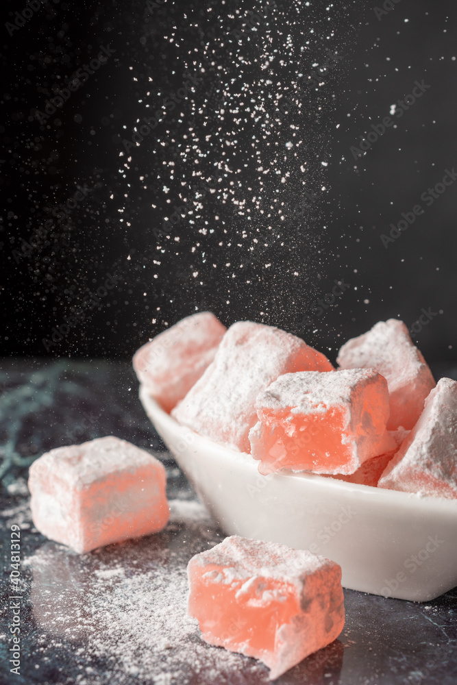 Fototapeta premium Top view of Turkish Delight with roses in white bowl, with sugar falling on blue marble, selective focus and black background, vertical