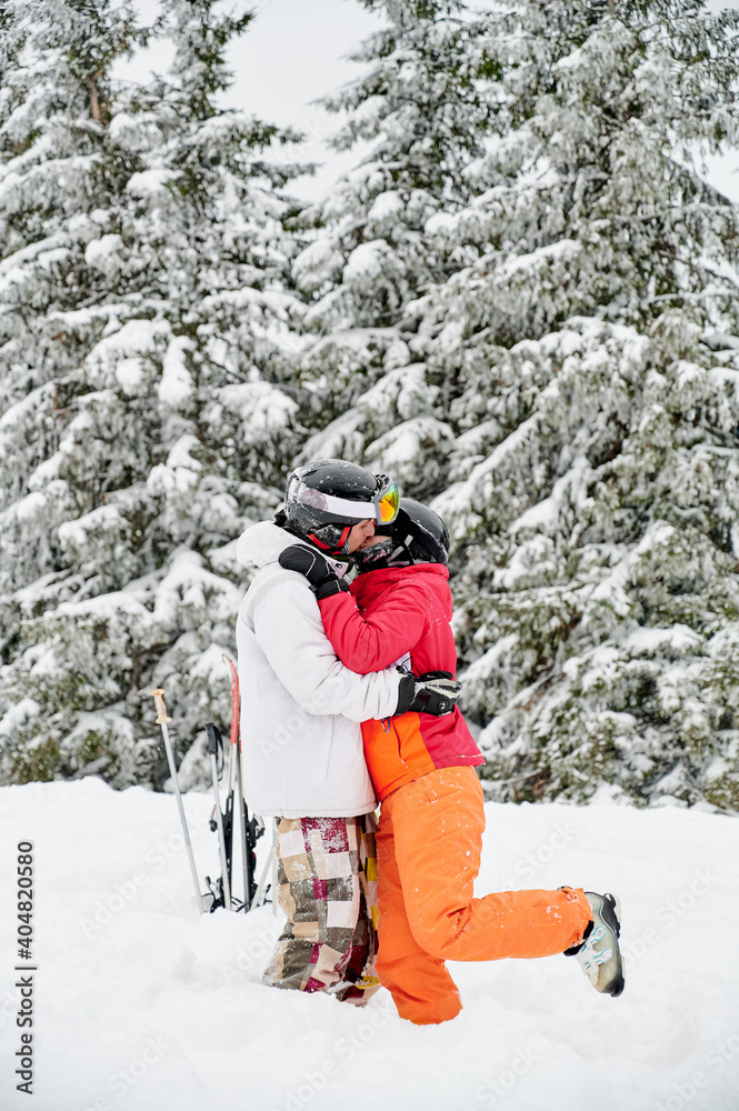 Young couple of tourists kissing in snow against mesmerizing snowy spruce forest on background. Vivid colorful suits on white snow. Concept of skiing and relationships.