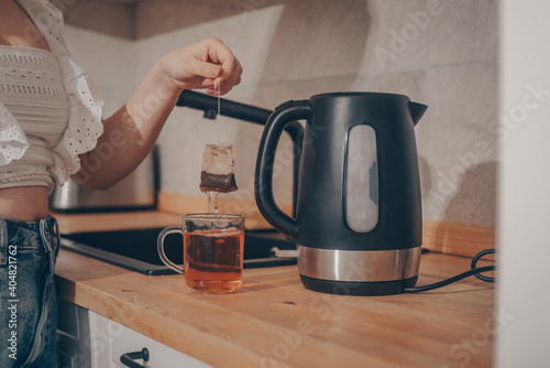A black kettle is on the kitchen table. A woman's hand holds a teapot and makes tea. Tea bag in a mug with water, tea leaves.