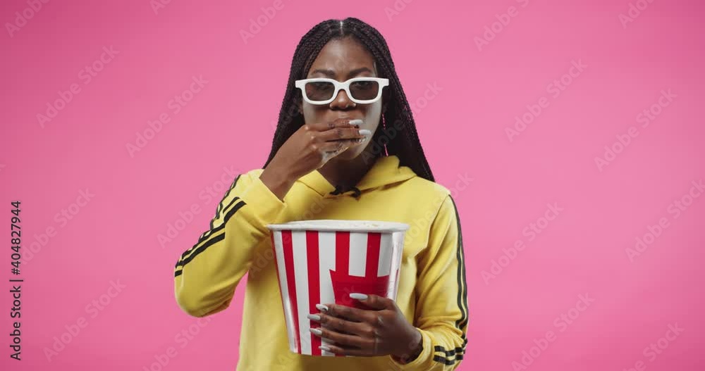 Portrait of African American young pretty happy woman holding popcorn box isolated on pink background. Female in yellow hoodie eating popcorn and watching movie in glasses. Woman watch film
