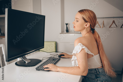 A beautiful girl looks into a large monitor and prints something. Woman works from home, freelance. A beautiful blonde looks at the computer screen.