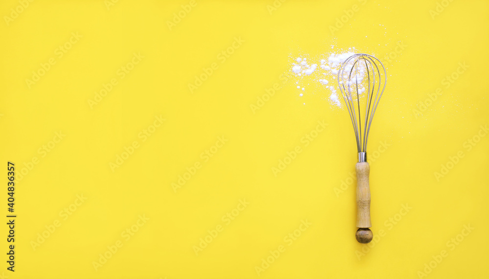 Obraz premium Whisk with wooden handle and white wheat flour on yellow background. Cooking utensil, egg beater mixer whisker . Flat lay banner background.