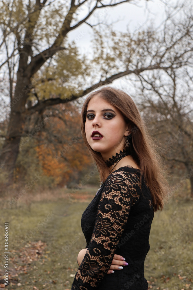 The girl in a black dress stands in the garden. Brown-haired woman with dark Gothic make-up: dark lips and eyes. Around the autumn weather. The woman has a black choker on her neck.