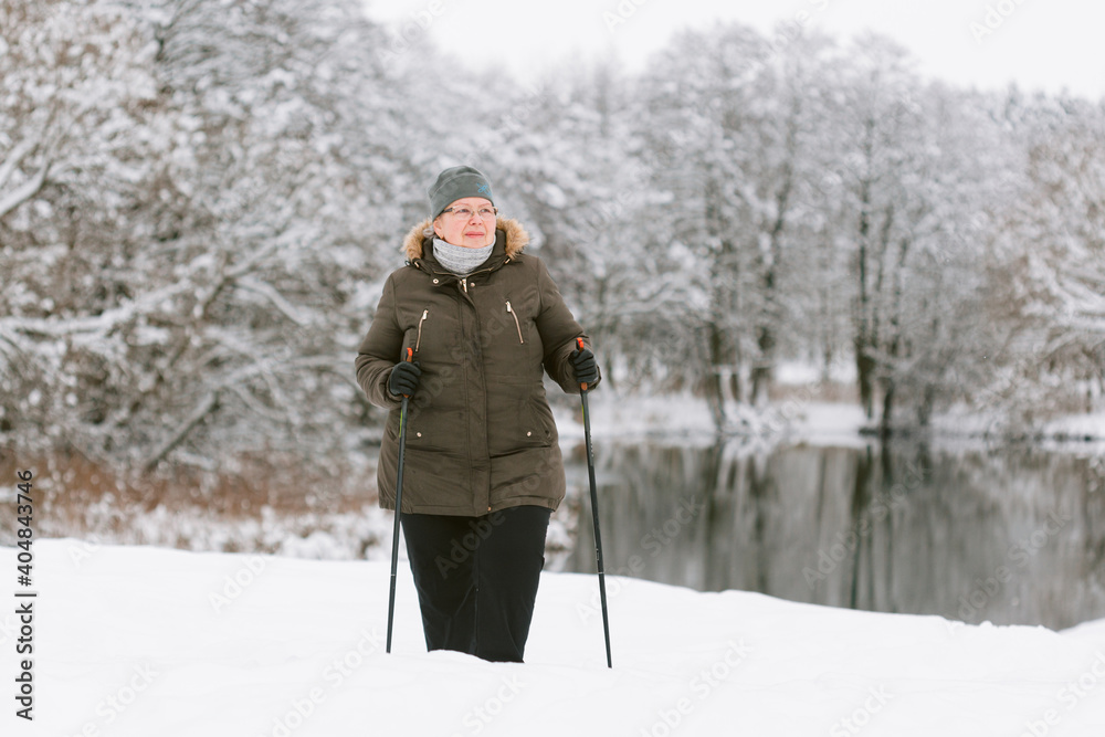 Senior woman standing with nordic walking poles in winter  park. Healthy lifestyle concept. Mature woman resting after exercise outdoors.