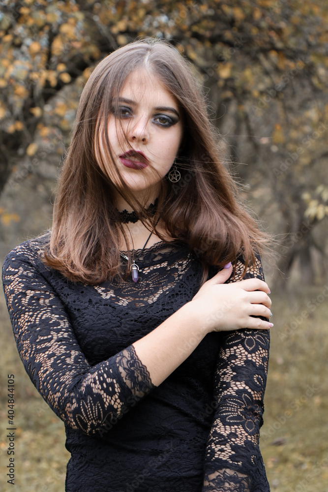 A young witch poses in the autumn forest. Golden tones around. The girl is dressed in a black dress and accessories in the Gothic style. Halloween shooting.