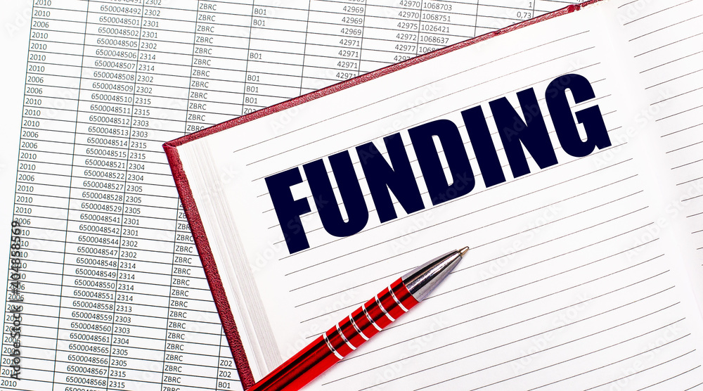 On the table next to the reports is a diary with the text FUNDING. Nearby is a red handle. On the table next to the reports is a diary with the text FUNDING. Nearby is a red handle.