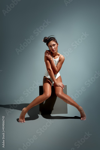 Sensual woman with long dark hair in white underwear sitting on gray cube over light grey studio background