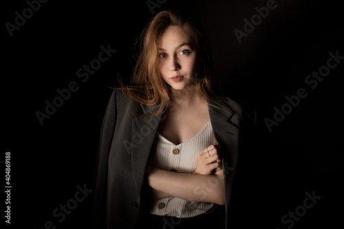 Young woman in jacket and white t-shirt standing alone in dark room and looking at camera, black background. Studio shot in low key. Casual portrait of girl with natural makeup and clean skin.
