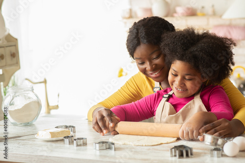 Happy black mom and her daughter rolling up dough together in kitchen