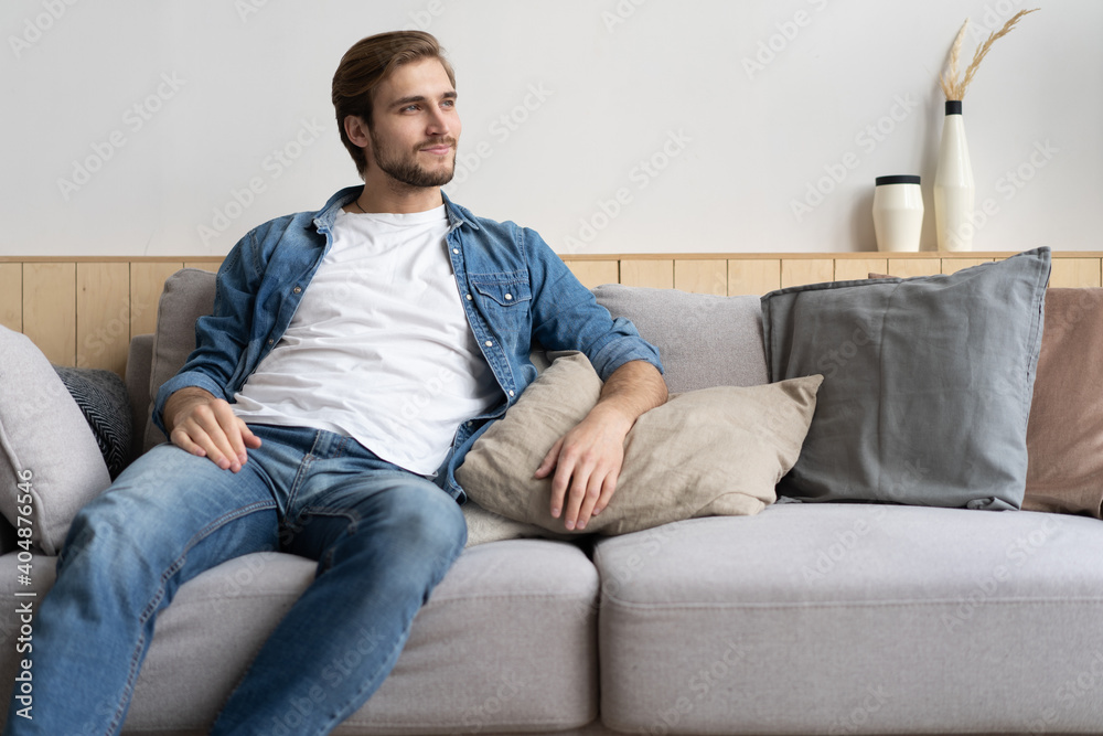Handsome young man looking thoughtful while sitting on the couch at home Handsome young man looking thoughtful while sitting on the couch at home