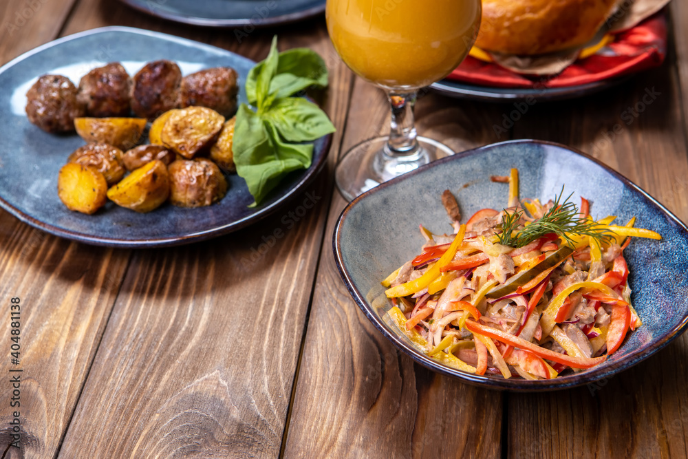 A cup with a salad of vegetables seasoned with sauce, fresh pastries, orange juice and fried potatoes with cutlets on a brown wooden background. A cup with a salad of vegetables seasoned with sauce, fresh pastries, orange juice and fried potatoes with cutlets on a brown wooden background.