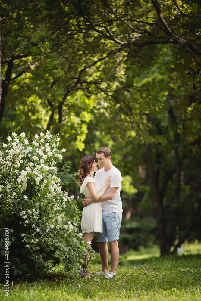 Young man and woman couple in a blooming garden near jasmine bush. Tender holding each other. Spring love story. Brown-haired girl with long hairs with her boyfriend or husband. Young couple on a date