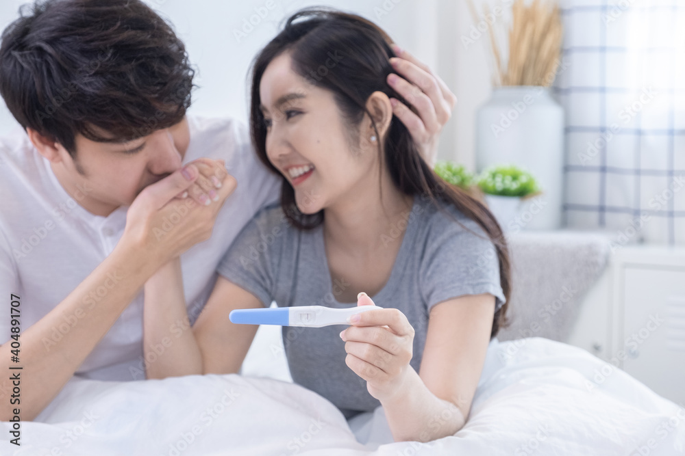 Happiness young married Asian couple with wife holding pregnancy test in hand and husband kissing her hand in bedroom focus on pregnancy test. Happiness young married Asian couple with wife holding pregnancy test in hand and husband kissing her hand in bedroom focus on pregnancy test.