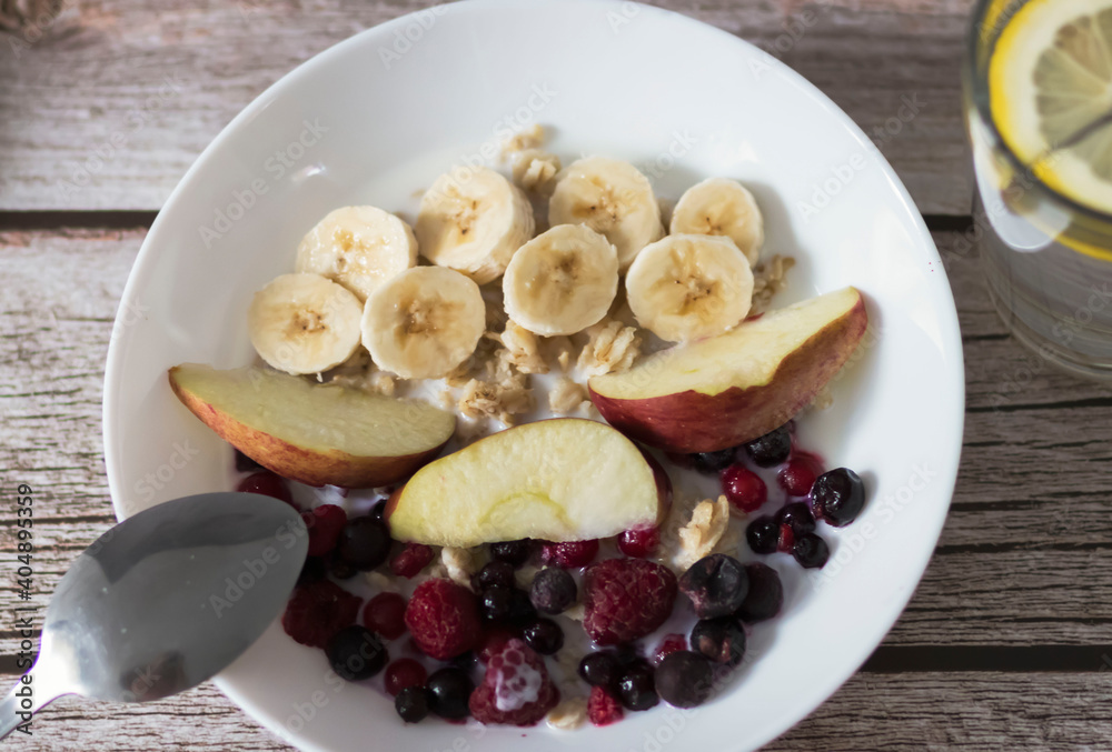 Oatmeal with banana pieces and berries, drenched in milk. Next to a glass of water with lemon