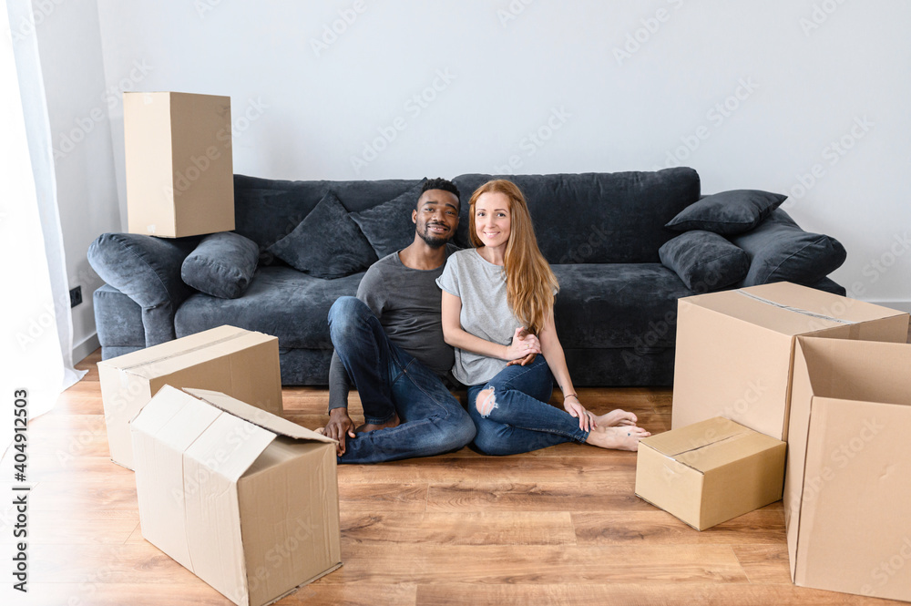Married mixed race couple in new own house. Smiling newlyweds sit on floor in living room, around cardboard boxes. Caucasian woman and african american man unpacking stuff, happy about relocation Married mixed race couple in new own house. Smiling newlyweds sit on floor in living room, around cardboard boxes. Caucasian woman and african american man unpacking stuff, happy about relocation