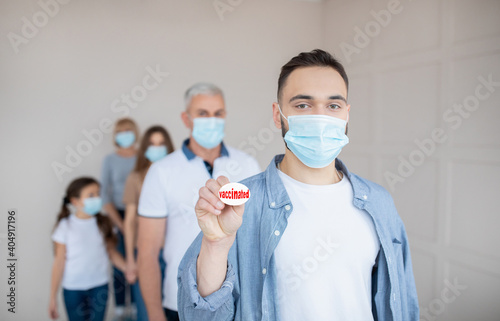 Young guy in protective mask holding VACCINATED badge in queue for coronavirus immunization at clinic, blank space