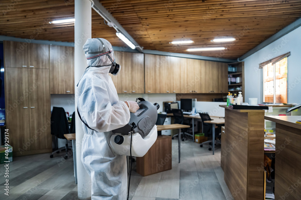 Professional technical man in prevention suit with his sterilizing machine and disinfecting water sprays all areas in the office room to take purifying coronavirus (COVID-19).